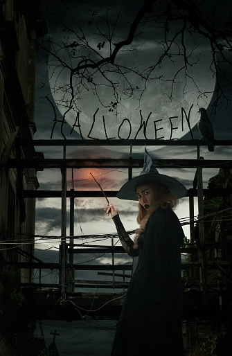 Halloween witch holding magic wand standing over damaged old wooden bridge, bird, dead tree, full moon with spooky cloudy sky, Halloween mystery concept