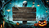 istock Halloween Party Card - Pumpkins And Skeleton In Graveyard At Night With Wooden Board 1338907781