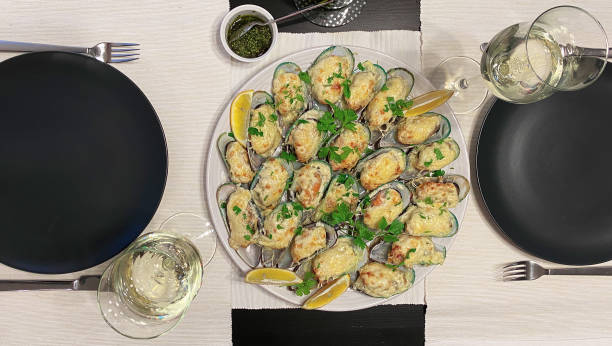halves of New Zealand green Perna canaliculus mussels baked with cream sauce and cheese on a platter, decorated with lemon wedges. Two glasses of white wine and black plates stock photo