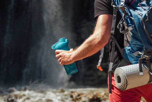 Hand close up with drinking water bottle. Man with backpack dressed in active trekking clothes touristic staying near mountain river waterfall and enjoying Nature. Traveling, trekking concept image stock photo