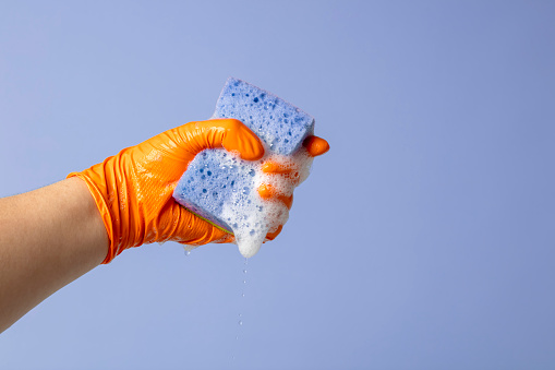 Close-up of a hand holding a wet cleaning sponge with soap sud. Blue background. Space for copy.