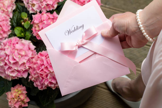 A hand holding a wedding invitation A female hand holding a wedding invitation in a pink envelope. Pink flowers in the background and part of the woman's shoes and dress. wedding invitation stock pictures, royalty-free photos & images