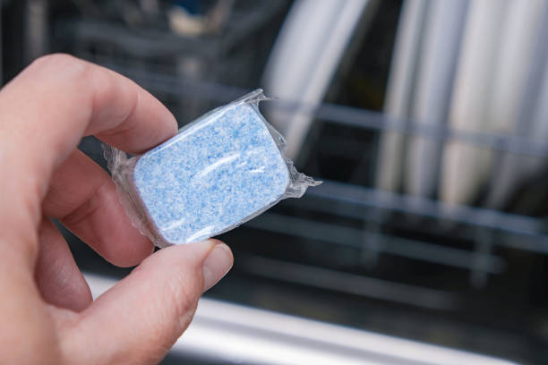 A hand holding blue dishwasher soap tablet in a water-soluble packaging on a dishashind machine background stock photo