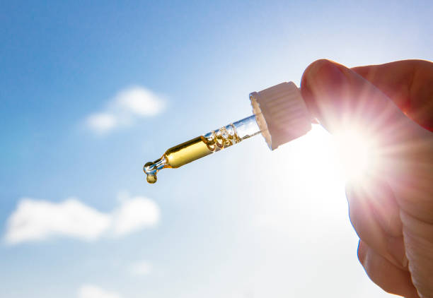 Hand holding dropper pipette with nice golden liquid D-vitamin against sun and blue sky on sunny day. Vitamin D keeps you healthy while lack of sun in winter, cure concept. stock photo