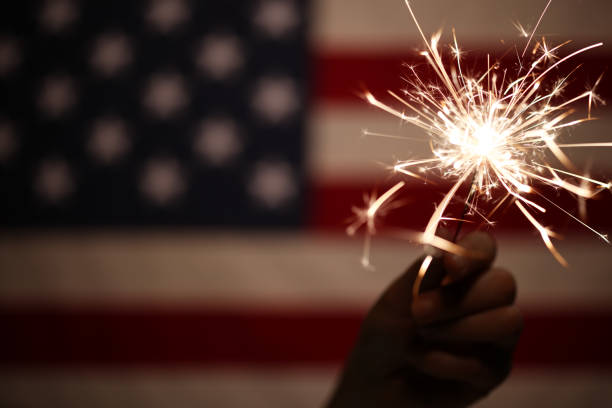 mano sosteniendo destello iluminado frente a la bandera americana para la celebración del 4 de julio - fourth of july fireworks fotografías e imágenes de stock