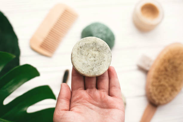 Hand holding natural solid shampoo bar on background of bamboo brush, deodorant, sponge on white wood with green monstera leaves. Zero waste. Choice plastic free eco products stock photo