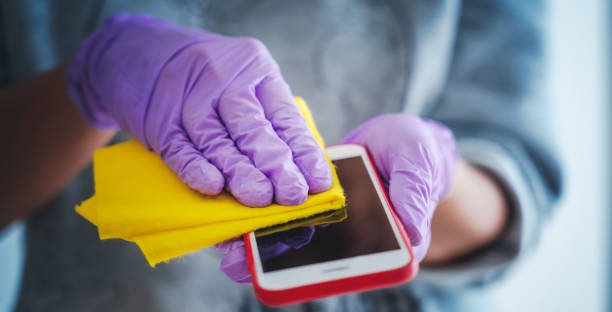 hand in purple glove wipes smartphone screen with a napkin, disinfection of surfaces stock photo
