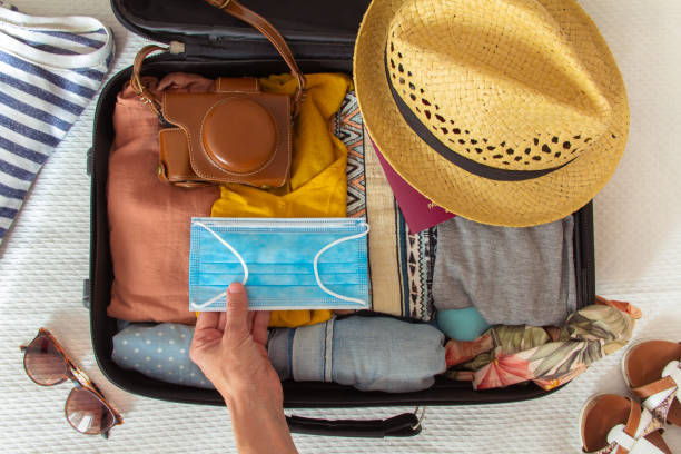 hand putting a face mask on the suitcase prepared to travel in summer, in the new normal, after the coronavirus covid 19 pandemic. there are other accesories like a hat, a camera and a bikini. - travel imagens e fotografias de stock