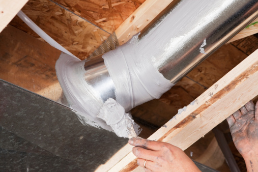 A dirty male hand, holding a paintbrush, is spreading caulk over a house air duct joint with a paintbrush. The caulk has been applied from a tube with a caulk gun and the brush is evenly spreading it over the joint.  Caulking residential ducts prevents air-conditioned or heated air from escaping the duct and prevents ambient air from entering the system. This is a new practice in homebuilding to increase energy efficiency, eventually it may be come standard or mandatory.