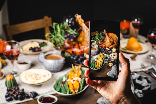 Person holding mobile phone and taking picture of thanksgiving meal served on dining table.