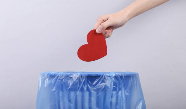 Hand throws red heart into trash bin with package on gray background stock photo