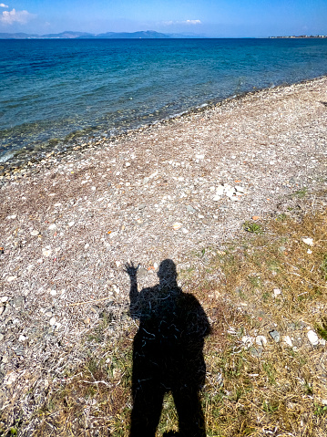 Summer vacation travel concept: Human shadow on beach ground. Hand waving gesture on a pebbly sea beach on a sunny day. Rest concept, lifestyle, relaxation, leisure, calm.