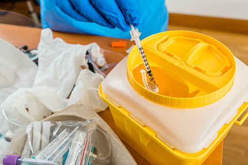 Hand with blue latex glove throws syringe into yellow waste container in hospital