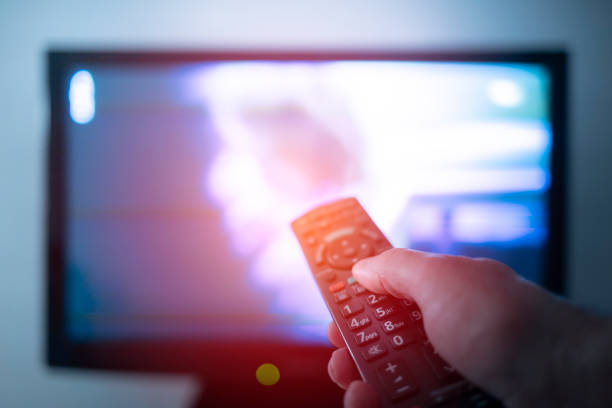 Hand with remote control in front of the tv What is on TV today? let's zap through all the channels television industry stock pictures, royalty-free photos & images