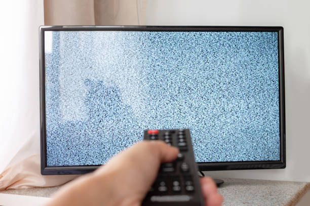 Hand with TV remote control in front of the screen with white noise on it - tuning the television channels and connecting problems stock photo