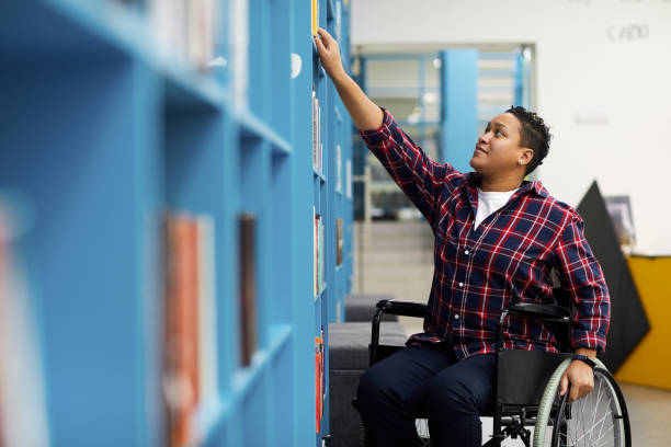 Handicapped Student in Library Portrait of disabled student in wheelchair choosing books while studying in college library, copy space physical disability stock pictures, royalty-free photos & images