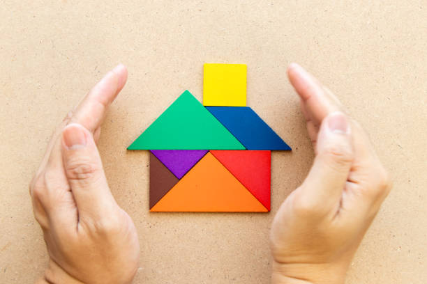 las manos sostienen el rompecabezas del tangram en la forma casera (concepto para el hogar, la protección de la familia o el seguro) - tangram casa fotografías e imágenes de stock