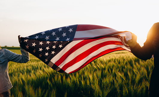 Hands holding American flag in a wheat field at sunset. Independence Day, 4th of July