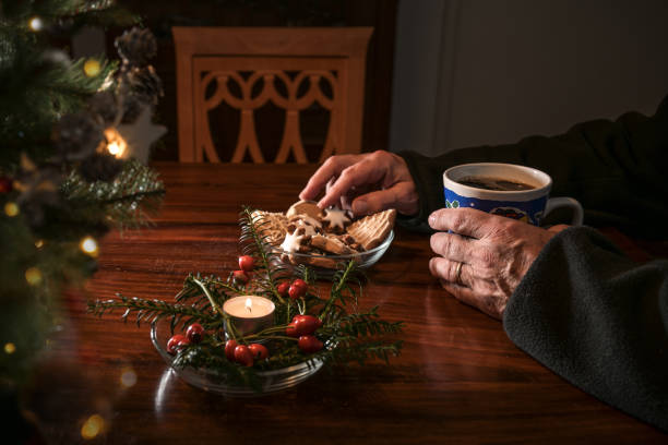Hands of an elderly single man sitting alone at a table with Christmas cookies, coffee and festive decoration next to an empty chair, lonely holidays during the croronavirus pandemic or after a loss stock photo