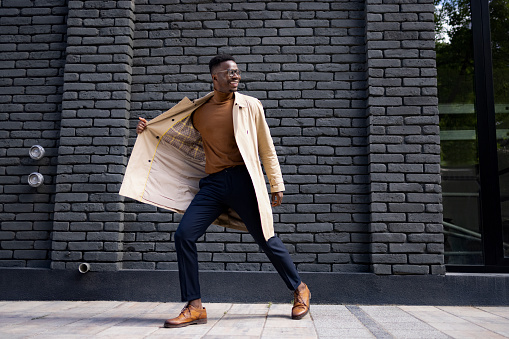 Attractive and handsome African American male model wearing a fashionable coat posing while standing on a city street and smiling
