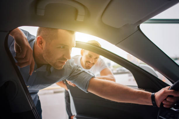 Handsome and confident customer stands and leans to car. He holds steering wheel with one hand. Guy is attentive to details. Manager stands behind him and lean to car. He is smiling and watching. stock photo