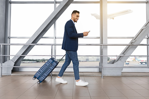Handsome arab guy using smartphone while walking with suitcase at airport terminal, young middle eastern man browsing mobile internet on cellphone while going to flight boarding, side view