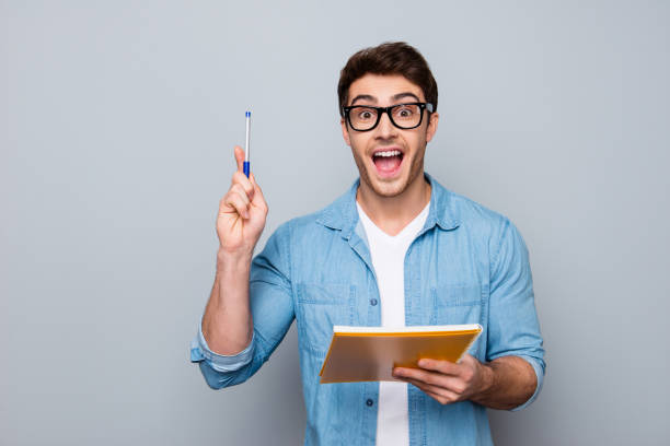 handsome, attractive, glad, positive, funny guy in glasses with wide open mouth finally found a solution how to make exercise, having raised pen and notebook in hands, isolated on grey background - man with pen imagens e fotografias de stock