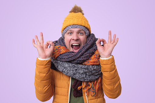 Handsome caucasian shocked funny man in several hats and scarfs showing ok gesture on colored background. Winter fashion.