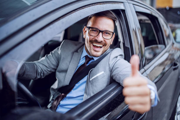 Handsome caucasian unshaven classy businessman in suit and with eyeglasses holding hand on steering wheel and giving thumbs up.  man driving suit stock pictures, royalty-free photos & images