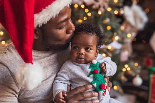 An attractive father of African American descent gives his newborn daughter a kiss on the forehead. The dad is sitting in front of the Christmas tree and wearing a Christmas hat.