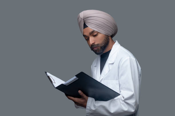 Handsome focused male doctor scrutinizing his patient files Waist-up portrait of a concentrated practitioner staring at medical documents in the open folder turban bebe fille stock pictures, royalty-free photos & images