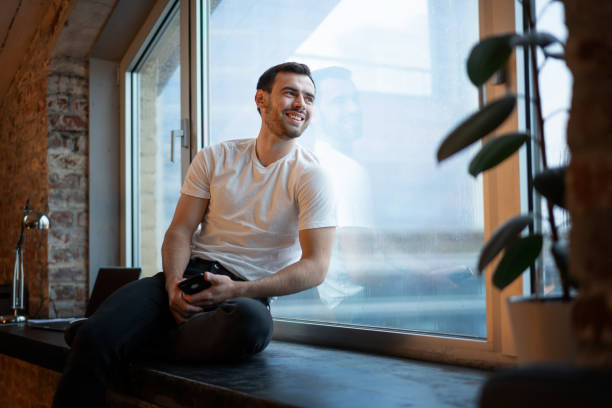 Handsome happy young brunette man in white t-shirt sitting on the windowsill at home on a rainy day with a phone in  hands stock photo