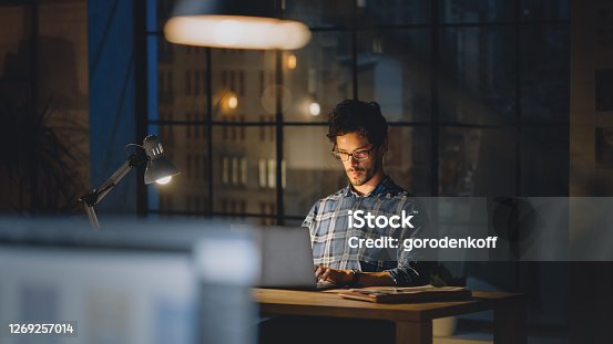 istock Handsome Male Creative Office Employee Sitting at His Desk Works on the Laptop. Evening in the Stylish Office Studio with Big Cityscape Window 1269257014