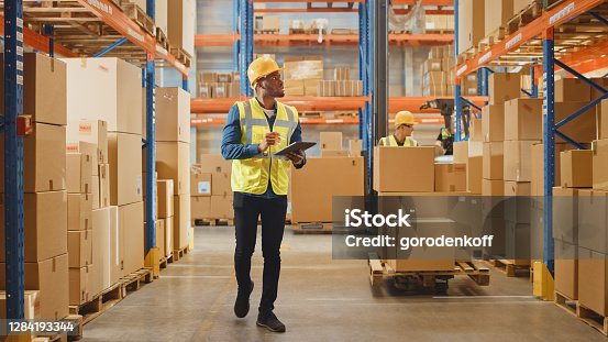 istock Handsome Male Worker Wearing Hard Hat Holding Digital Tablet Computer Walking Through Retail Warehouse full of Shelves with Goods. Working in Logistics and Distribution Center. 1284193344