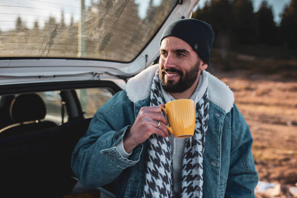Hombre guapo tomando café mientras está sentado en un automóvil en un día frío de invierno Hombre guapo tomando café mientras está sentado en un automóvil en un día frío de invierno hombres chaquetas de invierno fotografías e imágenes de stock