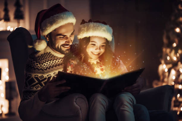 Handsome man is sitting in chair with his little cute daughter. Merry Christmas and Happy New Year! Handsome man is sitting in chair with his little cute daughter in Santa Claus hats and reading book at home. Magic light is coming from the inside. christmas story telling stock pictures, royalty-free photos & images