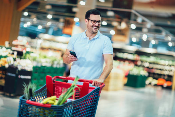 Handsome man shopping in supermarket pushing trolley and holding phone. stock photo