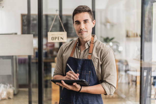 Handsome man standing holding a tablet and taking notes, he's a store clerk, he wears an apron and serves the customers who come to use the service in the cafe. Cafe service concept. stock photo