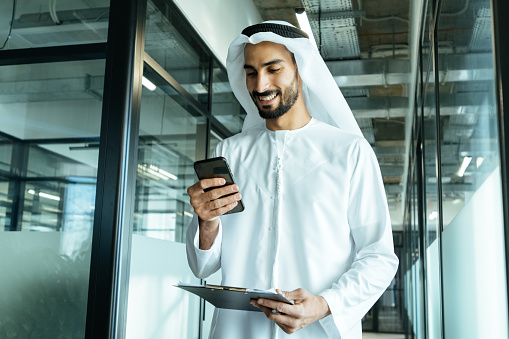 handsome man with dish dasha working in his business office of Dubai. Portraits of a successful businessman in traditional emirates white dress. Concept about middle eastern cultures, lifestyle and professional occupations