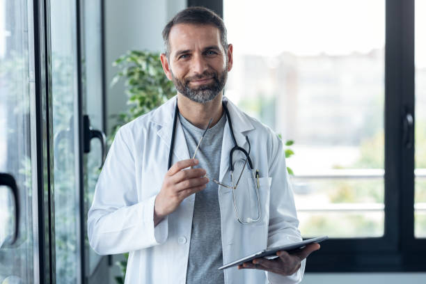 Handsome mature male doctor workinh while holding digital tablet looking at camera in the medical consultation. stock photo