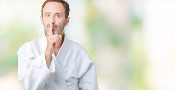 Handsome middle age senior man wearing kimono uniform over isolated background asking to be quiet with finger on lips. Silence and secret concept. Handsome middle age senior man wearing kimono uniform over isolated background asking to be quiet with finger on lips. Silence and secret concept. how do you say shut up in japanese stock pictures, royalty-free photos & images
