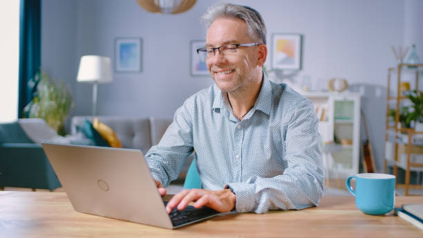 handsome middle aged man verwendet laptop-computer, während er an seinem schreibtisch im gemütlichen und stilvollen wohnzimmer sitzt. happy successful man arbeitet auf dem computer von zu hause aus. - einzelner mann über 40 stock-fotos und bilder