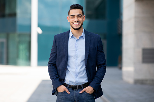 Handsome middle-eastern guy young businessman in stylish suit posing next to office center, smiling arab entrepreneur having break after successful busienss meeting, standing on street, copy space
