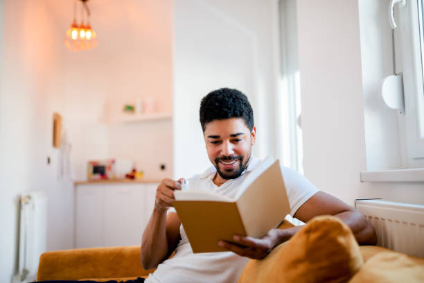 handsome smiling man reading a book while sitting on the sofa. - ler imagens e fotografias de stock