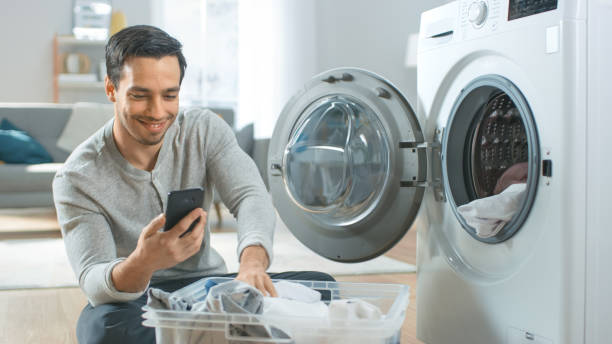 knappe glimlachende jonge mens in grijze jeans en de laag zit voor een wasmachine en gebruikt zijn smartphone. hij laadt washer met dirty laundry. lichte en ruime woonkamer met modern interieur. - washing machine stockfoto's en -beelden