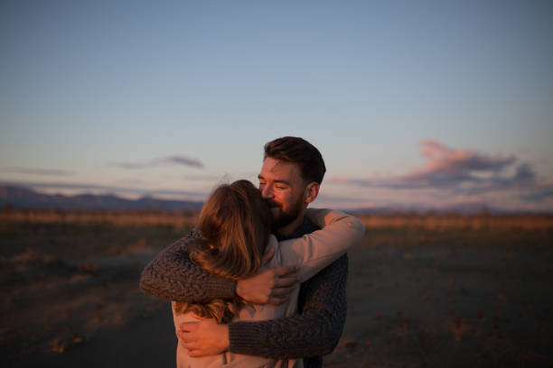 Handsome young bearded man hugging his girlfriend and watching at the sunset from the beach in the winter time stock photo