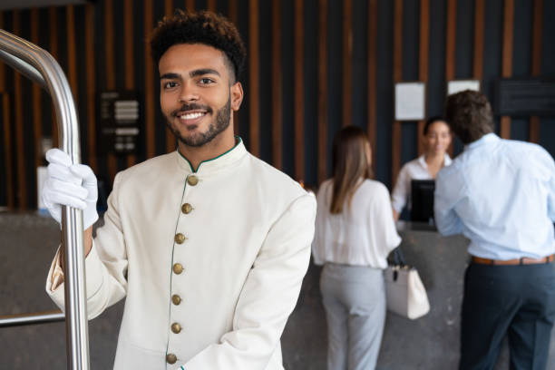 Handsome young black bellhop looking at camera smiling while holding luggage cart Handsome young black bellhop looking at camera smiling very happy while holding luggage cart hotel reception photos stock pictures, royalty-free photos & images