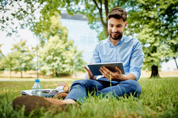 Handsome young businessman sitting outdoors in the park and using tablet stock photo