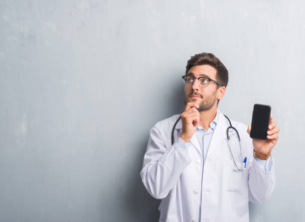 handsome young doctor man over grey grunge wall holding smartphone serious face thinking about question, very confused idea - doctor wall imagens e fotografias de stock
