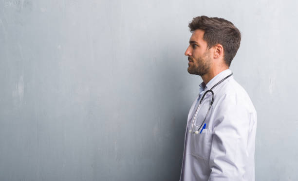 handsome young doctor man over grey grunge wall looking to side, relax profile pose with natural face with confident smile. - doctor wall imagens e fotografias de stock